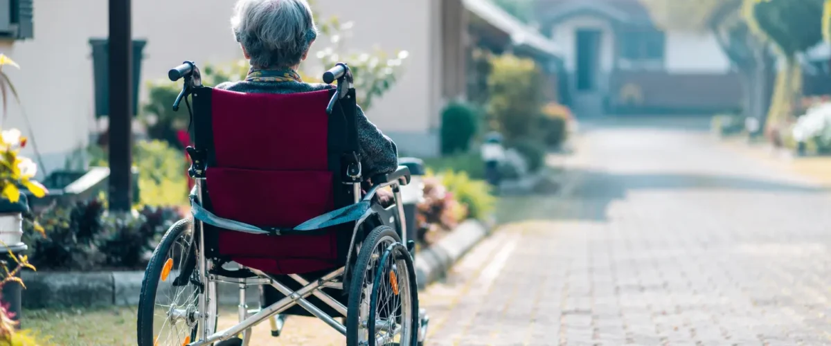 Anziana residente in una casa di riposo in Italia nel 2026 seduta su una sedia a rotelle in un tranquillo giardino della struttura, con vialetto curato, aree verdi e ambiente accogliente per l’assistenza agli anziani.