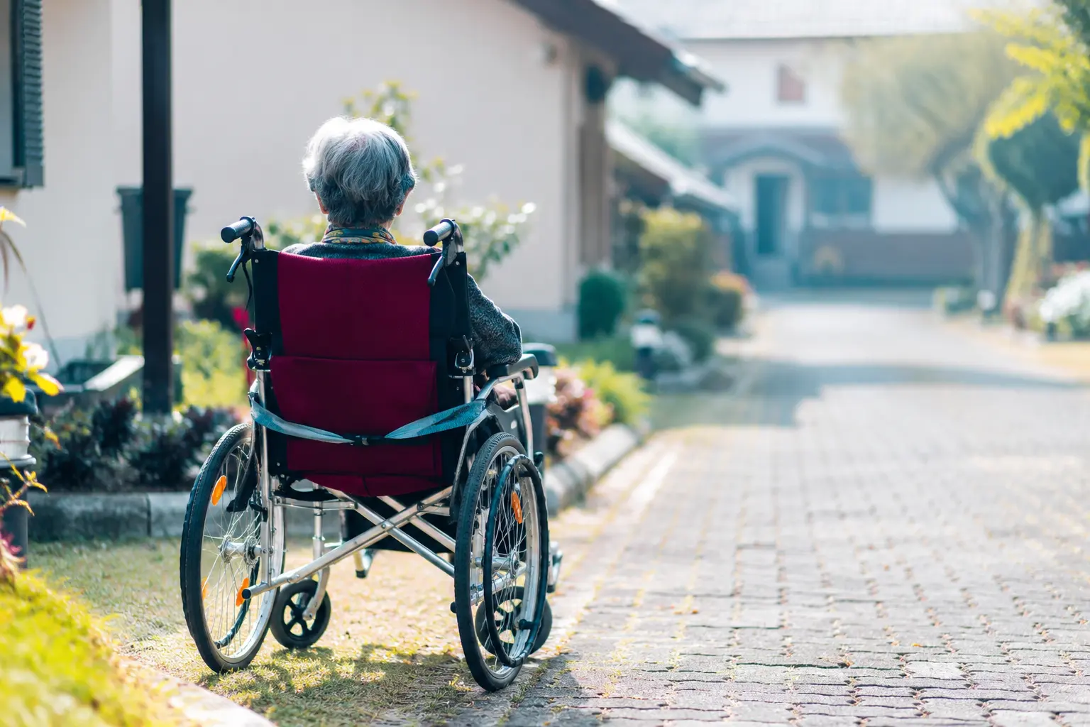 Anziana residente in una casa di riposo in Italia nel 2026 seduta su una sedia a rotelle in un tranquillo giardino della struttura, con vialetto curato, aree verdi e ambiente accogliente per l’assistenza agli anziani.